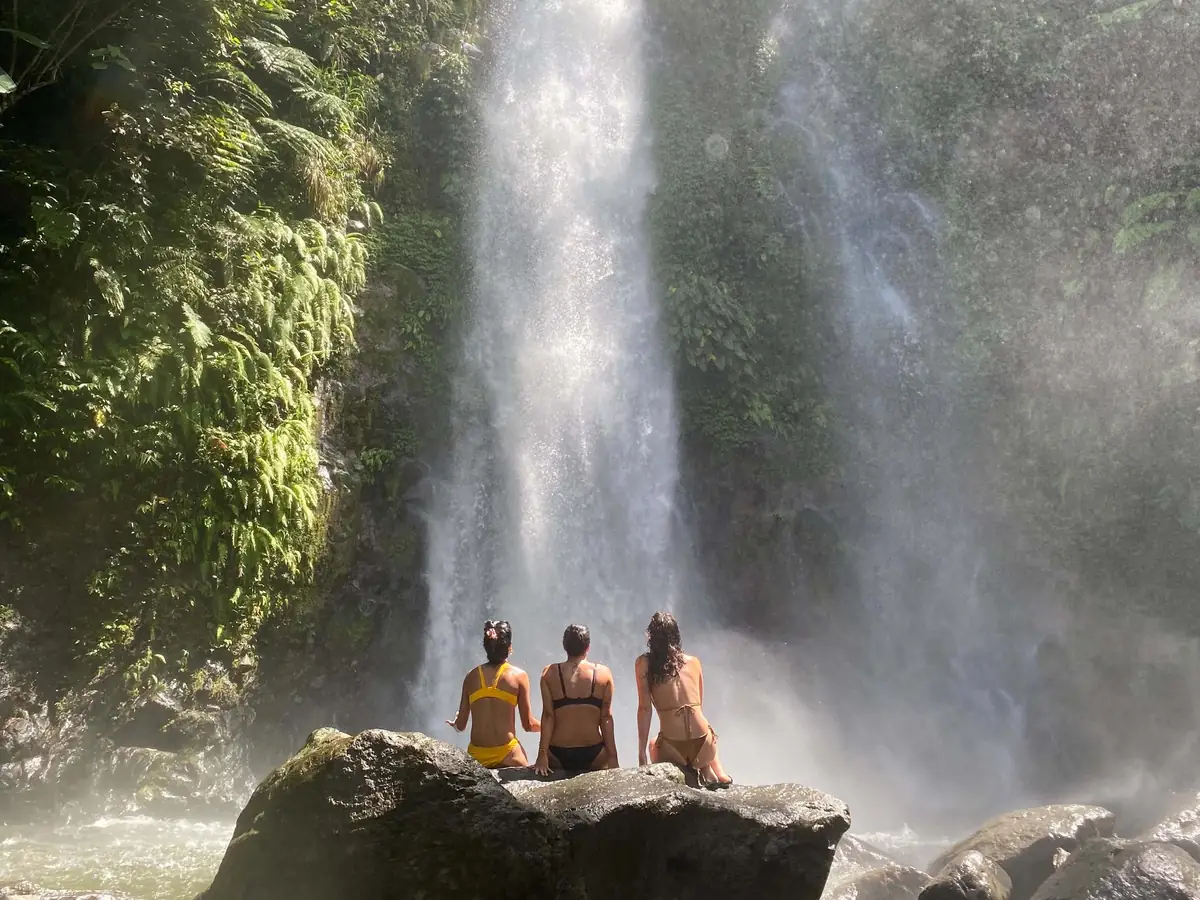 Three guests sitting on a boulder in the spray of a tall waterfall on Biliran Island