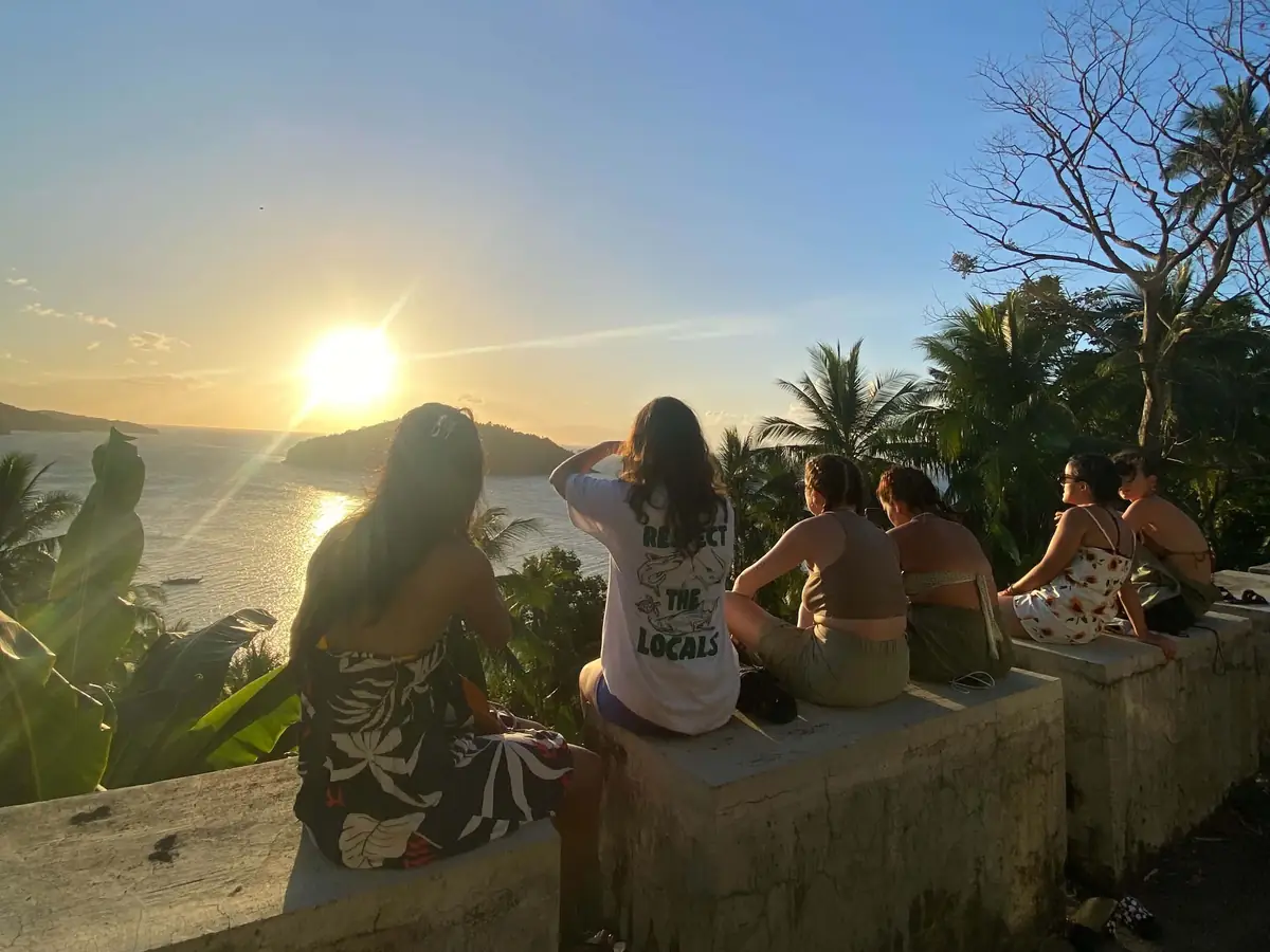 A row of guests sitting on a hilltop wall as the sun sets over the bay