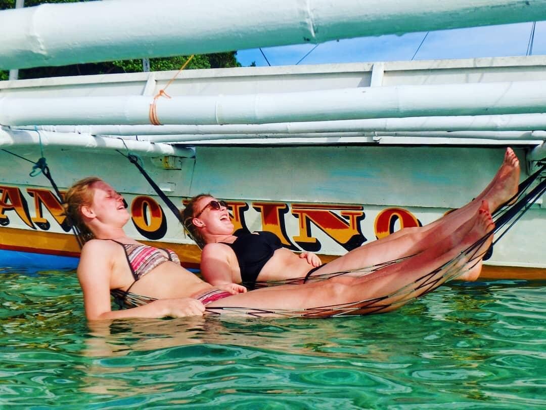 Two guests floating and relaxing beside a traditional paraw sailboat