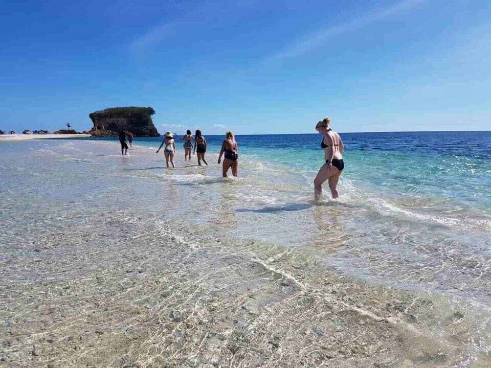 Travellers wading through clear shallow water near a rock islet