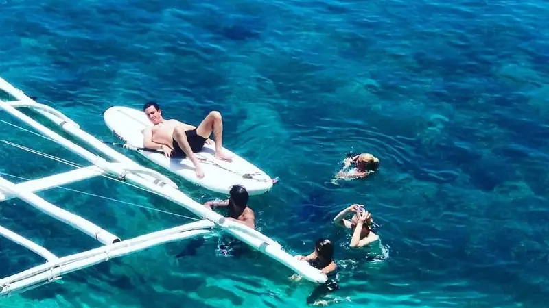 Guests swimming beside the bangka's outrigger in clear blue water off Lamanoc Island, one lying on a paddleboard