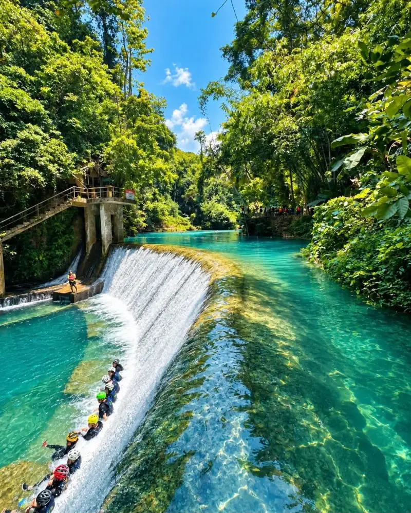 Canyoneers in helmets and life vests lined up along a waterfall weir above a turquoise river in the jungle, Badian, Cebu