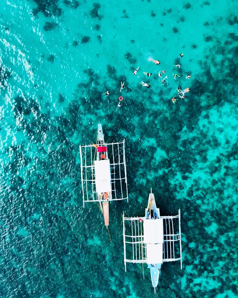 Drone view of two outrigger boats moored over a turquoise reef with snorkellers nearby, Malapascua, Cebu
