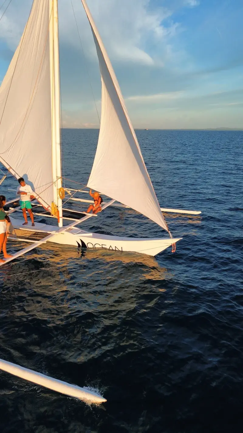 Drone view of guests on the deck of a white-sailed paraw on deep blue water