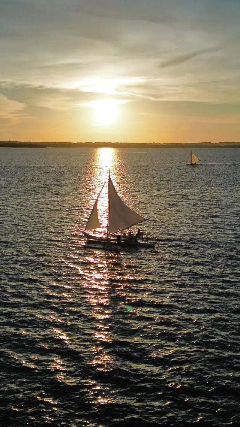 A paraw sailing far off on a calm sea beneath a low sun