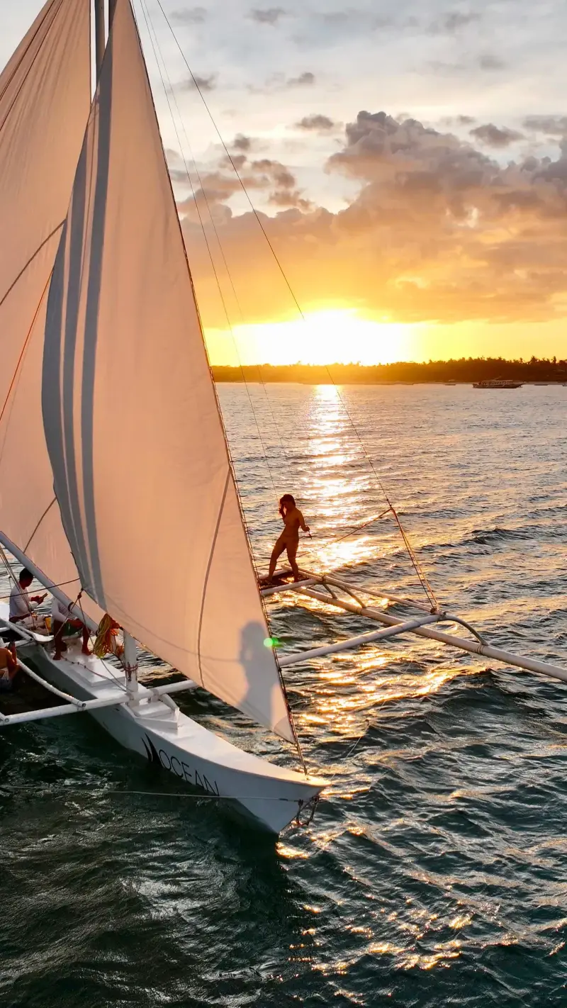 A paraw heeling under full sail at sunset, a guest standing on the outrigger
